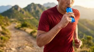 Un coureur de trail en t-shirt rouge consomme un gel énergétique bleu en courant sur un sentier de montagne.